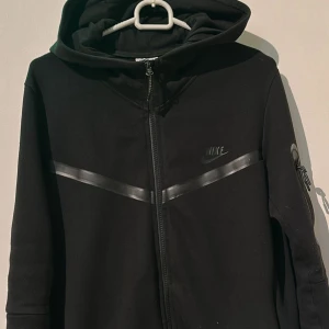 Svart hoodiejacka från Nike - Svart hoodiejacka från Nike med dragkedja och huva. Jackan har diskreta svarta detaljer och en diagonal rand över bröstet. Perfekt för en sportig stil och enkel att matcha med det mesta.