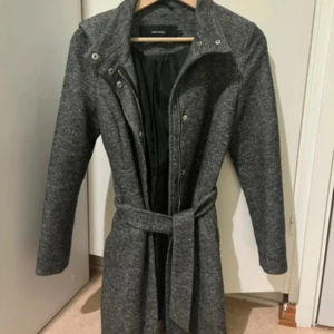 Grå kappa med bälte från Vero Moda - Säljer en stilren grå kappa från Vero Moda med bälte i midjan och knappar framtill. Jackan har lång ärm och en klassisk krage. Perfekt för dig som vill ha en enkel men snygg look.