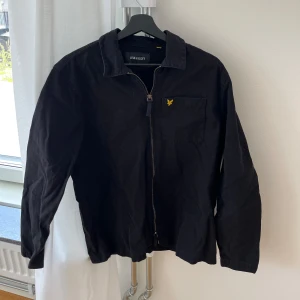 Svart jacka från Lyle & Scott - Svart jacka från Lyle & Scott med dragkedja framtill och klassisk krage. Bröstficka med gul broderad logotyp. Långärmad modell i bomull, perfekt för lager-på-lager-stil.