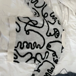 Grå stickad tröja med svart mönster - Säljer en grå stickad tröja med svart abstrakt mönster. Tröjan har långa ärmar och breda ribbade muddar vid ärmslut, nederkant och halsringning. Perfekt för dig som gillar unika och mysiga plagg.