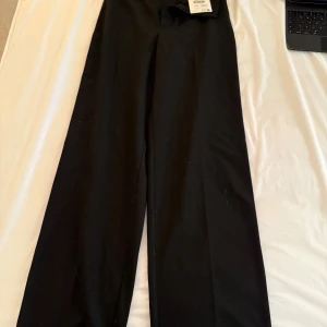Svarta kostymbyxor med vida ben - Säljer ett par svarta kostymbyxor med vida ben och hög midja. Byxorna har klassisk passform, bälteshällor och en bakficka. Perfekta för en stilren look och passar till många olika outfits.