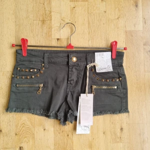 Gröna mini jeansshorts med nitar från Denim Co. - Snygga gröna jeansshorts från Denim Co. med råa kanter, guldfärgade nitar och dragkedjedetaljer framtill. Klassisk femficksmodell och knappstängning. Perfekta för en cool och avslappnad stil.