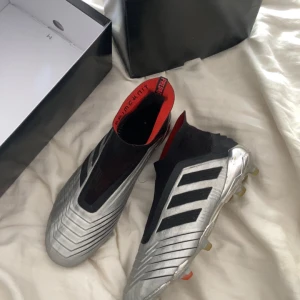 Adidas Predator 19+ fg  - Säljer ett par Adidas Predator 19+ fotbollsskor i silver med svarta detaljer och röda inslag. Skorna har en strumpliknande passform utan snörning och är designade för spel på gräs. Ikoniska tre svarta ränder på sidan.