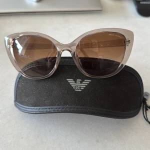 Bruna cat eye-solglasögon från Armani Exchange - Snygga solglasögon från Armani Exchange med transparent beige båge och bruna tonade glas. Cat eye-form ger en trendig look. Kommer med originalfodral med logga.