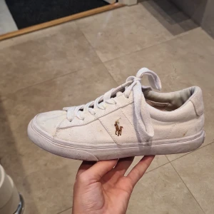 Vita sneakers från Polo Ralph Lauren - Säljer ett par vita sneakers från Polo Ralph Lauren med klassisk design och guldfärgad logga på sidan. Skorna har snörning och rund tå, tillverkade i textilmaterial med vit sula. Perfekta för en stilren vardagslook.