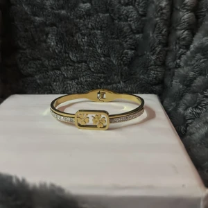 Guldfärgat armband med fyrklöver och stenar - Snyggt guldfärgat armband med rektangulär platta dekorerad med två fyrklöver. Armbandet har glittrande stenar längs sidorna och en elegant, smal design. Perfekt för att ge din outfit lite extra bling. Rostfritt!!