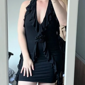 Svart volangklänning med djup v-ringning - Säljer en svart kortklänning med volangdetaljer och djup v-ringning. Klänningen är ärmlös och har öppen rygg, vilket ger en snygg och elegant look. Perfekt för dig som vill sticka ut med en stilren och trendig outfit.