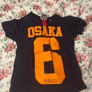 Mörkblå t-shirt Osaka 6 från Superdry - Mörkblå t-shirt från Superdry med stort gult tryck 'OSAKA 6' på framsidan och japanska tecken. Klassisk passform och korta ärmar. Perfekt för en sportig look eller streetstyle.