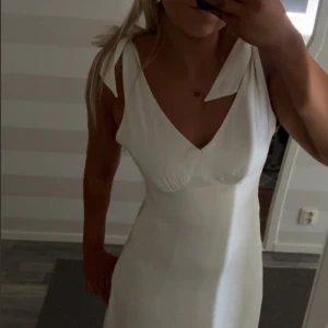 Vit långklänning med knytaxelband - Säljer en elegant vit långklänning med djup v-ringning och breda axelband som knyts på axlarna. Klänningen har öppen rygg och är figurnära upptill med en lätt utställd kjol. Perfekt för sommarens tillfällen!
