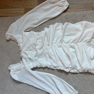 Vit långärmad omlottklänning med rynk - Säljer en vit långärmad klänning med omlottdetalj och rynk längs hela klänningen. Klänningen har puffiga ärmar med resår vid axlar och ärmslut. Använd en gång bara. 