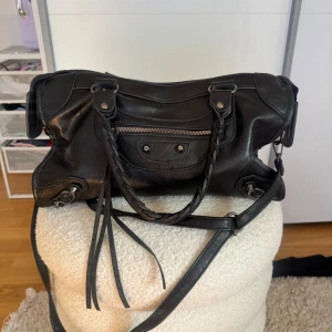 Svart axelväska med dragkedja och flätade handtag - Säljer en svart axelväska i syntetmaterial med både axelrem och två flätade handtag. Väskan har silverfärgade detaljer, dragkedja upptill och en liten ficka med dragkedja framtill. Perfekt för dig som vill ha en rymlig och stilren väska. ❗️Dragkedjan har gått sönder, se sista bilden❗️Den går att stänga men på ena sidan har den endast tappat tyg så den går att dra, den andra sidan har den lossnat från ena sidan. 