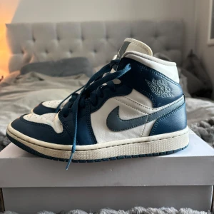 Nike Air Jordan 1 Mid blå och vita - Säljer ett par Nike Air Jordan 1 Mid i blått och vitt. Skorna har klassisk hög siluett, snörning och rund tå. Ovandelen är i skinn med blåa och vita paneler samt ikoniska Air Jordan-loggan på sidan.