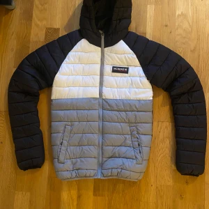 Svart och grå pufferjacka från McKenzie - Säljer en snygg pufferjacka från McKenzie i svart, vitt och grått. Jackan har huva, dragkedja framtill och två fickor med dragkedja. Perfekt för kyligare dagar och har en sportig look med McKenzie-logga på bröstet.