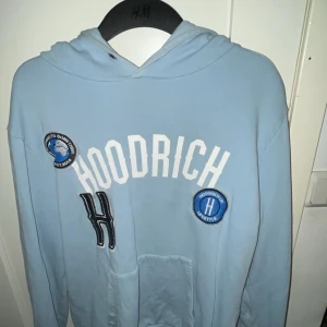 Ljusblå hoodie från Hoodrich - Säljer en ljusblå hoodie från Hoodrich med vita tryck och broderade patches på bröstet. Tröjan har huva och magficka, samt långa ärmar. Perfekt för en avslappnad stil.