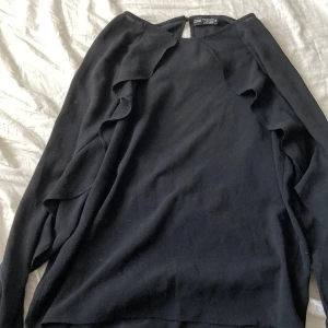 Svart långärmad topp med volangdetaljer från ONE TEE - Säljer en svart långärmad topp  med snygga volangdetaljer längs ärmarna. Toppen har en enkel rund halsringning och är tillverkad i ett mjukt material som känns skönt mot huden. Perfekt för dig som gillar stilrena plagg med en twist.