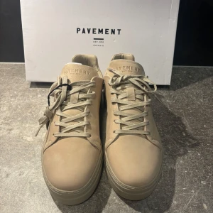 Beige sneakers från Pavement - Säljer ett par stilrena beige sneakers från Pavement med klassisk design och rund tå. Skorna har snörning och är tillverkade i skinn med platt sula. Perfekta för dig som gillar minimalistisk stil.