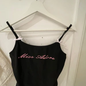 Svart linnetopp med text Miss Adore - Säljer en svart linnetopp med tunna axelband och rosa rosetter. Framtill står det 'Miss Adore' i rosa text. Perfekt för en trendig och söt look. Storlek S