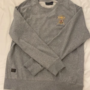 Grå sweatshirt från Morris R. Club - Säljer en klassisk grå sweatshirt från Morris R. Club med broderad logga och brittisk flagga på bröstet. Tröjan har rund halsringning och långa ärmar. Perfekt för en stilren och sportig look.