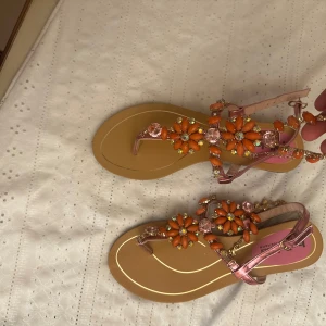 Blommiga sandaler med pärldetaljer - Säljer ett par sandaler från Bella med blommiga dekorationer i orange och rosa samt pärldetaljer. Remmarna är i metallicrosa och sandalerna har en platt sula. Perfekta för sommaren och ger en färgglad touch till din outfit!
