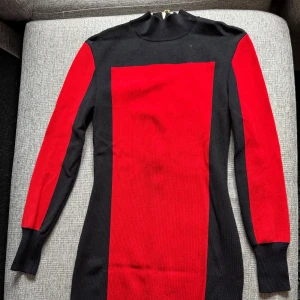 Svart och röd långärmad klänning från HM x Balmain - Säljer en snygg svart och röd klänning från HM x Balmain med lång ärm och hög krage. Klänningen har en tight passform och är tillverkad i ett stretchigt material som sitter skönt på kroppen. Perfekt för dig som vill sticka ut med färgblock!