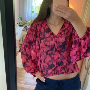 Rosa och svart blommig blus - Säljer en färgstark blus i rosa och svart med blommigt mönster. Blusen har långa, vida ärmar och knappar framtill. Modellen är croppad och har en lös passform, perfekt för dig som gillar statement-plagg. Storlek M men passar S