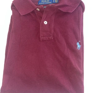 Vinröd pikétröja från Polo Ralph Lauren - Säljer en vinröd pikétröja från Polo Ralph Lauren med klassisk krage och två knappar framtill. Tröjan har blå broderad logga på bröstet och är i modellen custom slim fit. Perfekt för en stilren look.