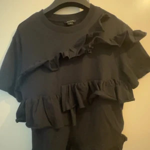Svart t-shirt med volanger från Monki - Svart t-shirt från Monki med två breda volangdetaljer framtill. Rund halsringning och korta ärmar. Perfekt för dig som vill ha en enkel topp med lite extra detaljer.