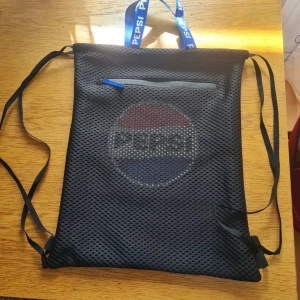 Ryggsäck med Pepsi-logga - Säljer en svart ryggsäck med nätmaterial och stor Pepsi-logga på framsidan. Väskan har blå dragkedja på ett ytterfack och blåa band med Pepsi-tryck. Kan hämtas i Vallentuna.