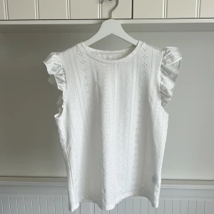 Vit ärmlös topp med volang - Säljer en söt vit topp med volangdetaljer på axlarna och broderat hålmönster. Toppen är ärmlös och har rund halsringning, perfekt för varma dagar eller sommarens alla tillfällen. Pris går att diskuteras