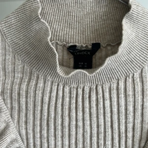 Beige ribbad tröja med volang från Lindex - Säljer en beige ribbad tröja från Lindex med långa ärmar och snygga volangdetaljer framtill. Tröjan har en hög, vågig krage och är perfekt för dig som gillar en trendig och bekväm stil. Priset går att diskutera 