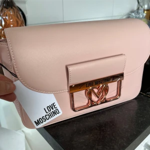 Ljusrosa axelväska från Love Moschino - Supersnygg ljusrosa axelväska från Love Moschino med guldfärgat spänne och logga framtill. Väskan har ett stilrent och elegant utseende med praktisk dragkedja och innerfack. Perfekt för att lyfta din outfit med en touch av lyx.