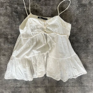 Vit broderad linnetopp från Zara - Säljer en söt vit linnetopp från Zara med broderade detaljer och tunna axelband. Toppen har volang nertill och en liten knytning framtill. 😍😍aldrig använd