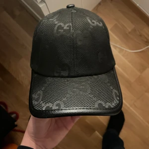 Svart keps från Gucci med GG-mönster - Snygg svart keps från Gucci med klassiskt GG-mönster över hela kepsen. Justerbar rem baktill i svart läder med präglad Gucci-logga. Kepsen är tillverkad i bomull och polyester, med detaljer i läder.