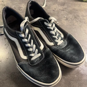 Svarta och vita Vans Old Skool sneakers - Klassiska Vans Old Skool sneakers i svart med vit sula och vit sidosnirkla. Skorna har snörning och är tillverkade i mocka och canvas. Perfekta för en avslappnad streetstil.