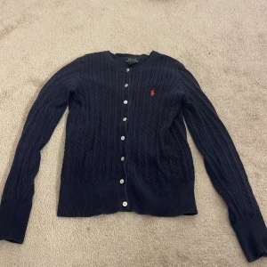 Mörkblå kabelstickad kofta från Polo Ralph Lauren - Säljer en mörkblå kabelstickad kofta från Polo Ralph Lauren med rosa logga (ser ut som röd i kameran). Använder inte längre. Pris kan diskuteras💕💕 