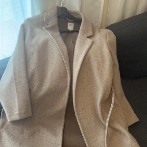 Beige kappa från ONLY - Säljer en stilren beige kappa från ONLY i storlek M. Kappan har klassisk krage, lång ärm och bälte i midjan för en snygg siluett. Perfekt för vår och höst. Kappan är använd 2 gånger 