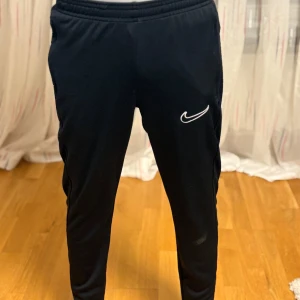 Svarta träningsbyxor från Nike - Svarta träningsbyxor från Nike med smal passform och elastisk midja. Byxorna har en vit Nike-logga på låret och dragkedjor vid fickorna. Perfekta för träning eller chill. Knappt använd.