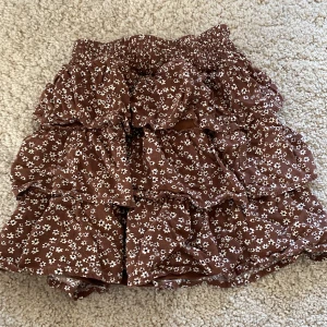 Brun blommig volangkjol från Lindex - Söt brun kjol med vita småblommor från Lindex. Kjolen har flera volanger och resår i midjan för en bekväm passform. Perfekt för dig som gillar en lekfull och trendig stil.