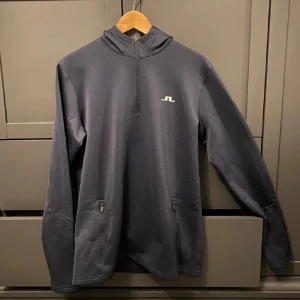 Mörkblå hoodie från J.Lindeberg - Säljer en mörkblå hoodie från J.Lindeberg med halv dragkedja och huva. Tröjan har långa ärmar, två fickor med dragkedja och en liten vit logga på bröstet. Perfekt för lager-på-lager och sportiga outfits.Nypris 1500!