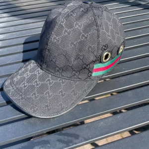 Svart keps från Gucci med monogram - Säljer en svart keps från Gucci med diskret GG-monogram över hela kepsen. Kepsen har två stora metallöljetter på sidan och ett grönt och rött band. Klassisk böjd skärm och justerbar passform.