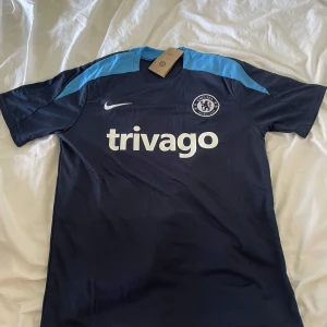 Chelsea FC tränings t-shirt från Nike - Mörkblå Chelsea FC tränings t-shirt från Nike med ljusblå detaljer på axlarna. Kortärmad modell med klubbmärke och Nike-logga på bröstet samt trivago som sponsortryck. Perfekt för fotbollsträning eller support.