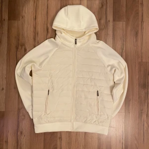 Vit quiltad hoodiejacka med dragkedja - Snygg vit hoodiejacka med quiltade partier och hel dragkedja framtill. Jackan har två fickor med dragkedja och en mysig huva. Perfekt för lager-på-lager och chill vardagsstil. Den är i L men passar M perfekt