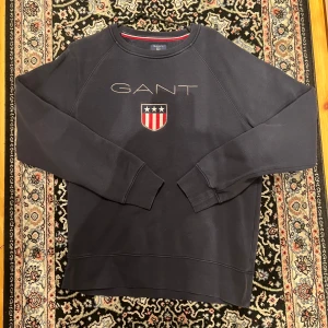 Mörkblå sweatshirt från GANT med broderad logga - Säljer en mörkblå sweatshirt från GANT med broderad logga och sköld med stjärnor och ränder på bröstet. Tröjan har rund halsringning och långa ärmar. Klassisk och stilren design med ribbade muddar.