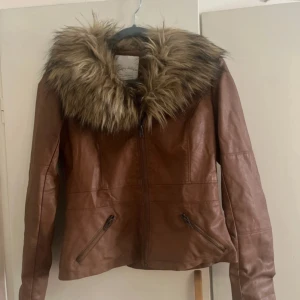 Brun skinnjacka med fuskpäls - Säljer en brun skinnjacka med stor, fluffig fuskpäls runt kragen. Jackan har dragkedja framtill och två snedställda fickor med dragkedja. Snygg och trendig modell som passar perfekt till hösten.