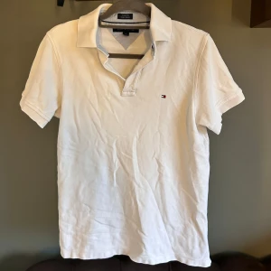 Vit piké t-shirt från Tommy Hilfiger - Vit piké t-shirt från Tommy Hilfiger med klassisk krage och korta ärmar. Liten broderad logga på bröstet i rött, vitt och blått. Perfekt för en clean och stilren look. Knappt använd.