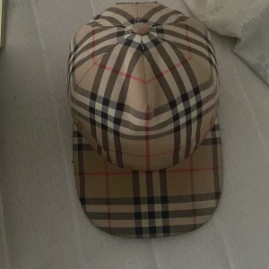 Burberry rutig keps - Säljer en klassisk rutig keps från Burberry i beige, svart och rött. Kepsen har justerbar baksida och ikoniskt Burberry-mönster. Märkeslogga bak. Perfekt accessoar för en trendig look. Det följer också med box och dushbag