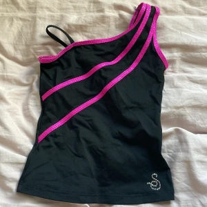 Svart one shoulder topp från Sagester med rosa paljettband - Snygg svart one shoulder topp från Sagester med glittriga rosa paljettband och strassdetalj nertill. Perfekt om man vill se lite extra piffig till konståknings träningen.
