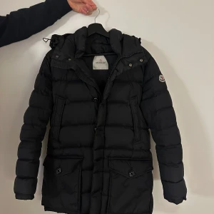 Svart dunjacka från Moncler - Säljer en svart dunjacka från Moncler med huva och stora fickor framtill. Jackan har pufferdesign, dragkedja och knappar samt Moncler-logga på ärmen. Perfekt för kalla dagar och riktigt snygg streetstil.