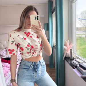 Vit blommig croppad topp med rosett Magda Butrym H&M - Superfin vit croppad topp med röda rosor och en stor rosett framtill. Toppen har korta ärmar och ett romantiskt blommönster. Perfekt för dig som vill sticka ut med en söt och trendig look.