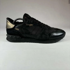 Valentino Rockrunner sneakers i svart och kamouflage - Säljer ett par Valentino Rockrunner sneakers i svart mocka och skinn med diskret kamouflagemönster och svarta nitar på hälen. Skorna har rund tå, platt sula och svarta skosnören. Perfekta för dig som gillar exklusiva och stilrena sneakers.